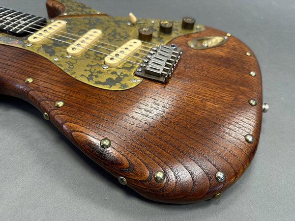Close-up of Coffee House Guitars electric guitar body made of polished wood with brass rivets and a unique brass pickguard with three cream pickups and metal bridge on gray background