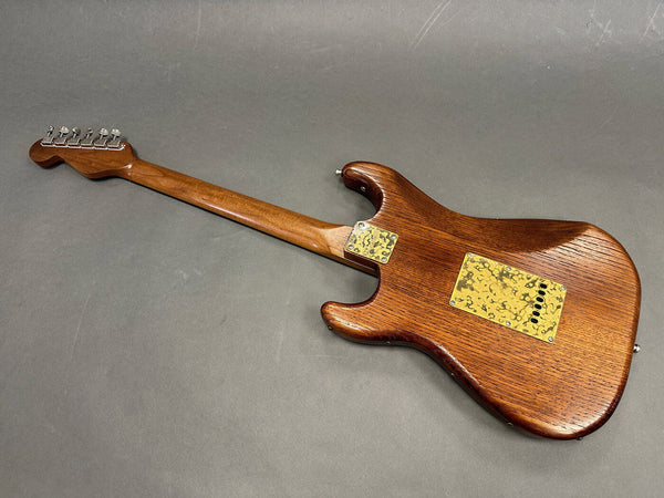 Back view of a wooden electric guitar with a dark brown finish and a vintage-style tin backplate on a gray surface