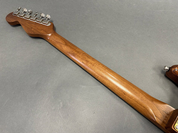 Close-up image of the back of a wooden electric guitar neck with chrome tuning pegs on a gray background