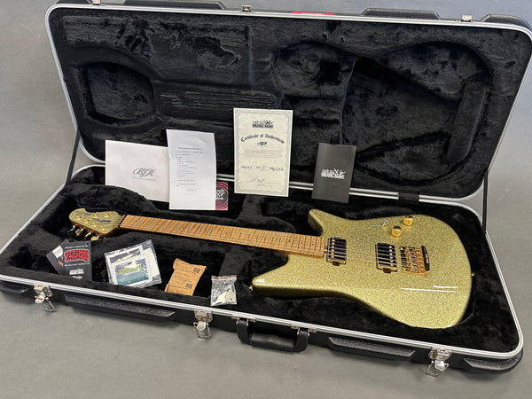 Ernie Ball Music Man electric guitar with gold sparkle finish and maple neck in black plush-lined hard case including certificate of authenticity, documentation, and guitar accessories.