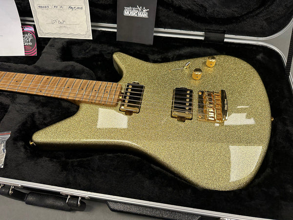 Ernie Ball Music Man electric guitar with gold sparkle finish, maple fretboard, dual humbucker pickups, and gold hardware in a black plush hard case