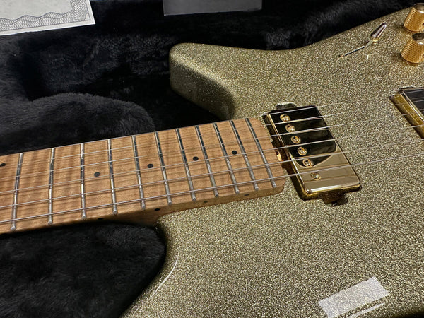 Close-up of electric guitar neck and body with gold sparkle finish, maple fretboard, gold humbucker pickup, and gold control knobs resting in a black plush guitar case.