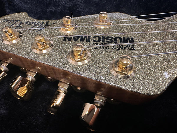 Close-up of Music Man guitar headstock with gold tuning pegs and sparkly finish on black background