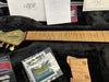 Ernie Ball Music Man guitar headstock with gold tuning pegs and maple fretboard in black plush-lined guitar case with maintenance accessories and inspection tags.