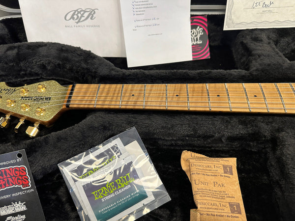 Ernie Ball Music Man guitar headstock with gold tuning pegs and maple fretboard in black plush-lined guitar case with maintenance accessories and inspection tags.