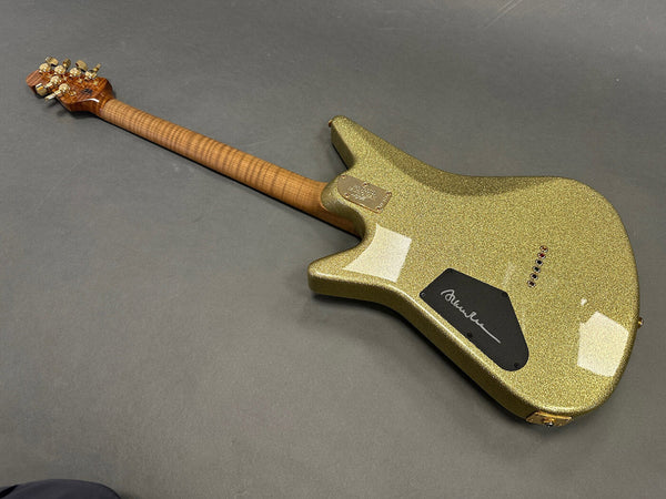 Back view of a gold sparkle electric guitar with a curly maple neck and gold tuning pegs on a gray background, featuring a black signature plate and six string ferrules.