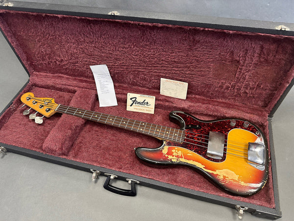 Vintage Fender Precision Bass guitar with sunburst finish and tortoiseshell pickguard in black hard case with red plush interior, includes Fender owner's manual and additional papers