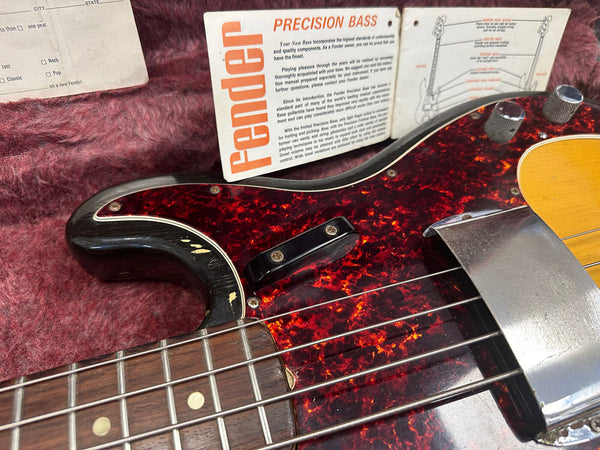 Close-up of vintage Fender Precision Bass guitar showing tortoiseshell pickguard, chrome pickup cover, control knobs, and rosewood fretboard with case lining and Fender Precision Bass instruction manual in background