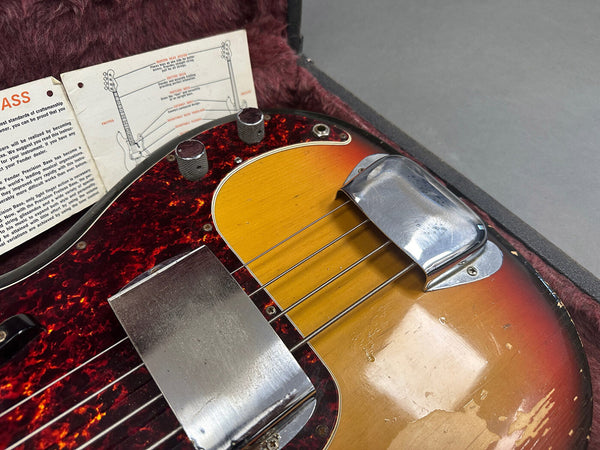Close-up view of vintage sunburst Fender Precision Bass with tortoiseshell pickguard and metal bridge and pickup covers, showing wear and included original Fender setup guide in plush-lined case.