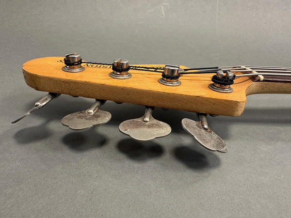 Close-up of a bass guitar headstock with four metal tuning pegs and wrapped strings against a gray background