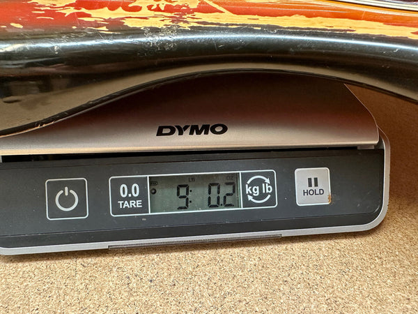 Close-up of a digital DYMO scale showing a guitar weight of 9 pounds 0.2 ounces, with part of the guitar body visible on top of the scale.