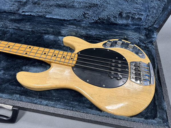 Natural wood finish electric bass guitar with black pickguard, chrome knobs, and maple neck resting in plush blue guitar case interior