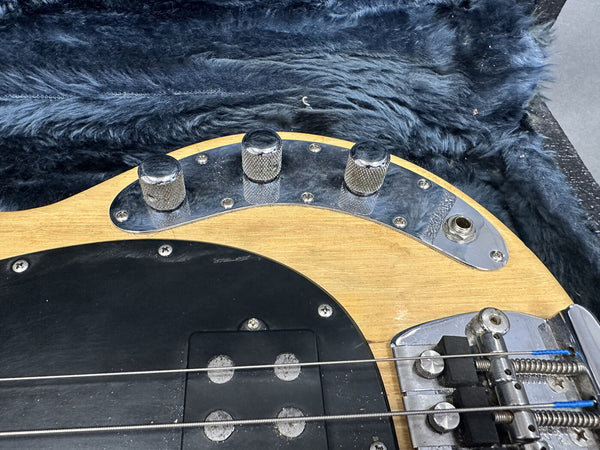 Close-up of natural wood electric bass guitar body featuring three chrome control knobs, output jack, black pickup cover, and bridge, resting on a black faux fur surface.
