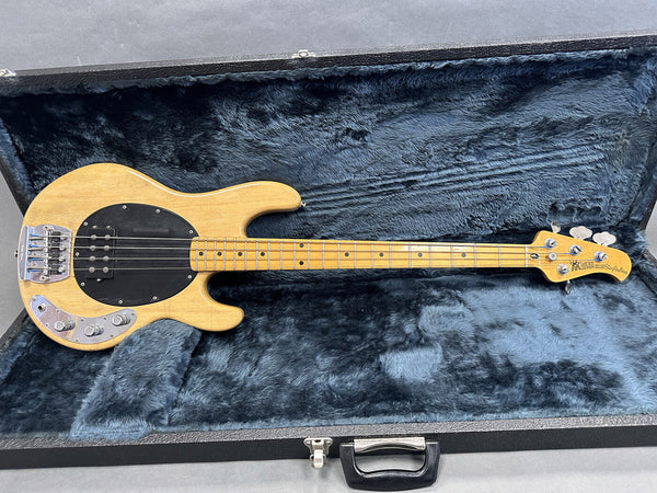 Natural wood finish Music Man StingRay 4-string bass guitar with maple fretboard and black pickguard, resting in a blue plush-lined hard case