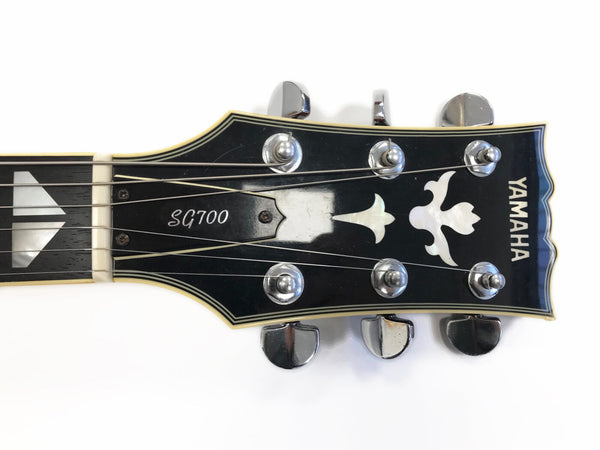Close-up of Yamaha SG700 electric guitar headstock with chrome tuning pegs and decorative inlay on black background